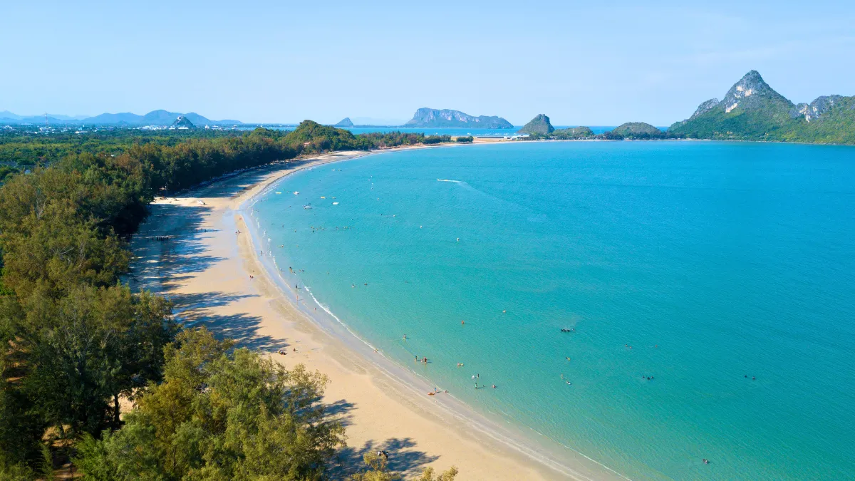 A curved tropical beach with turquoise water, trees, and distant islands under a clear sky