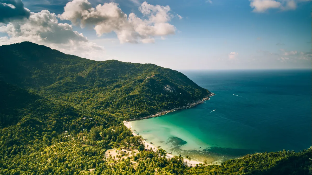 A tropical bay with green mountains, a sandy beach, and turquoise water under a partly cloudy sky