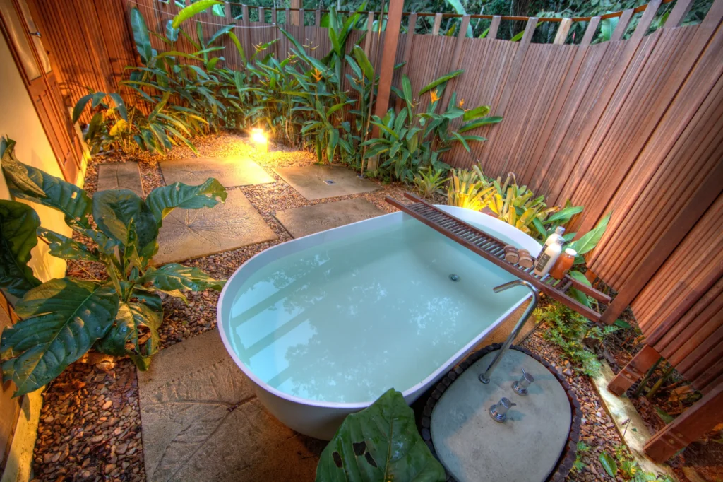 Outdoor soaking tub in a tropical garden courtyard with stone paths and wooden fencing