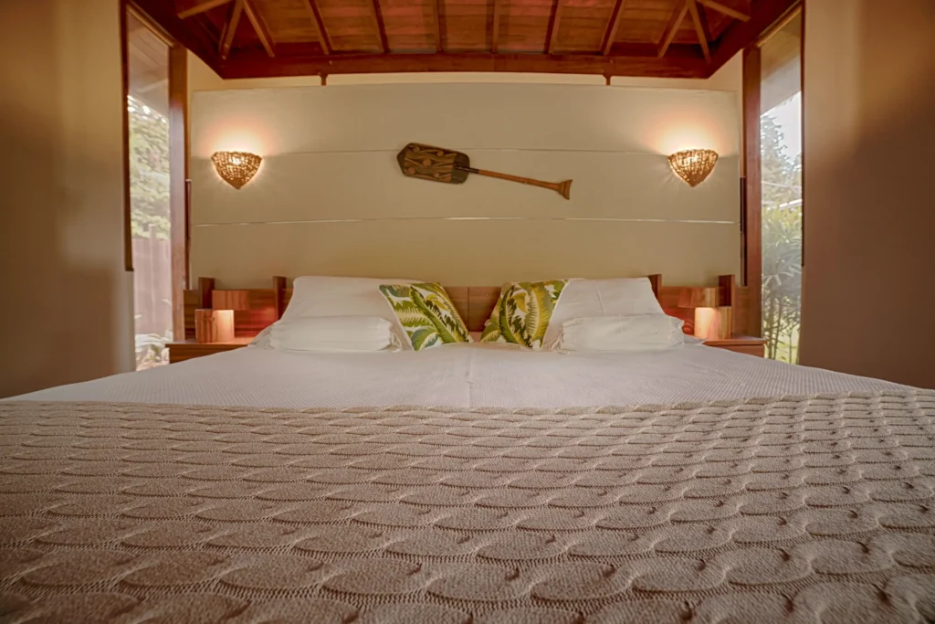 A cozy bedroom with a large bed, tropical pillows, and warm wall sconces under a wooden ceiling