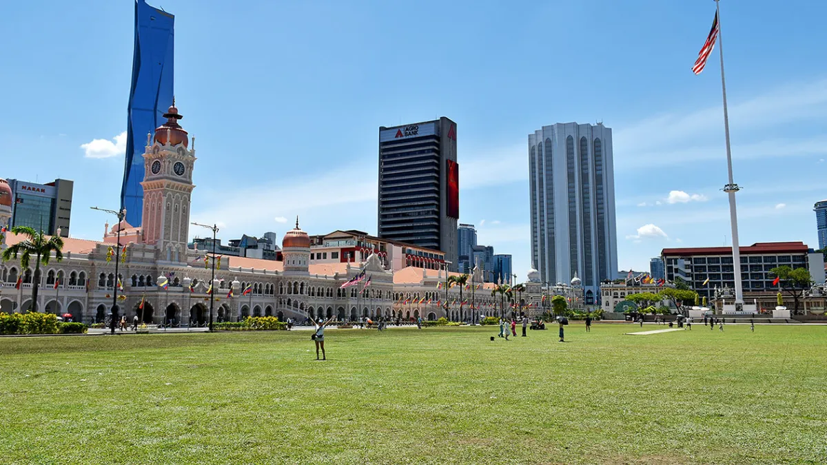 A large grassy field in front of historic colonial buildings and modern skyscrapers, with a tall flagpole and Malaysian flag
