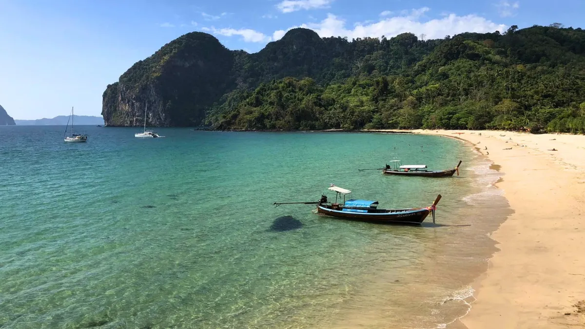 Tropical beach cove with clear turquoise water, longtail boats, and forested cliffs