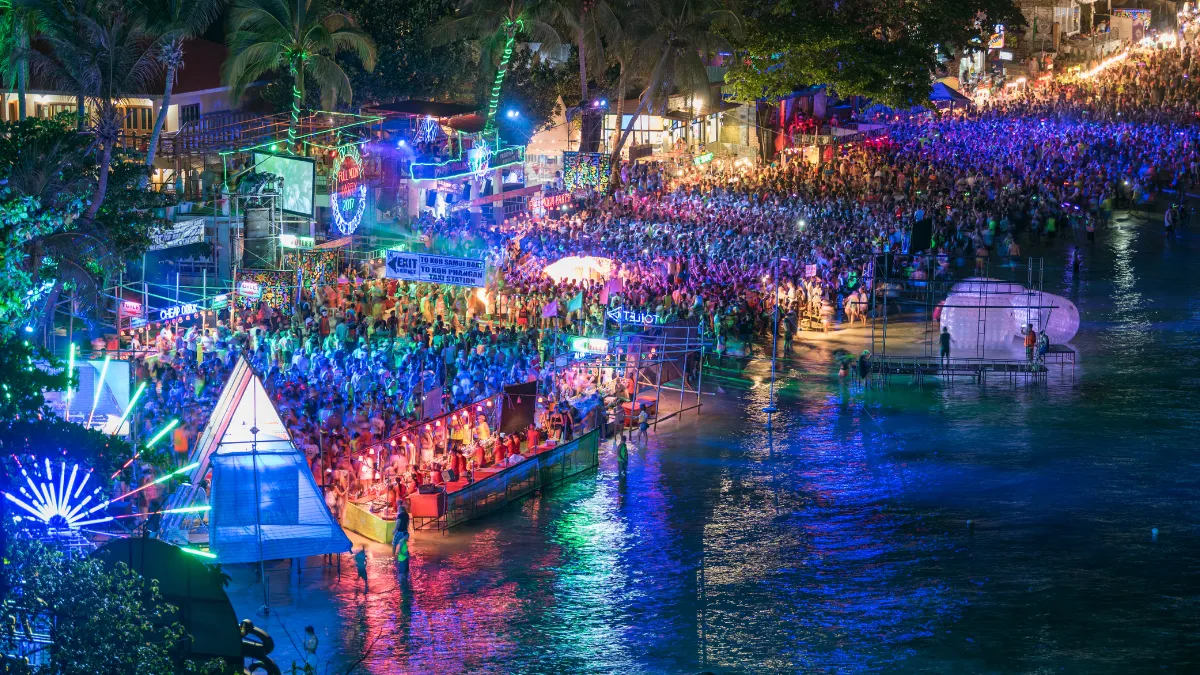 Crowded nighttime beach festival with colorful lights along the shoreline