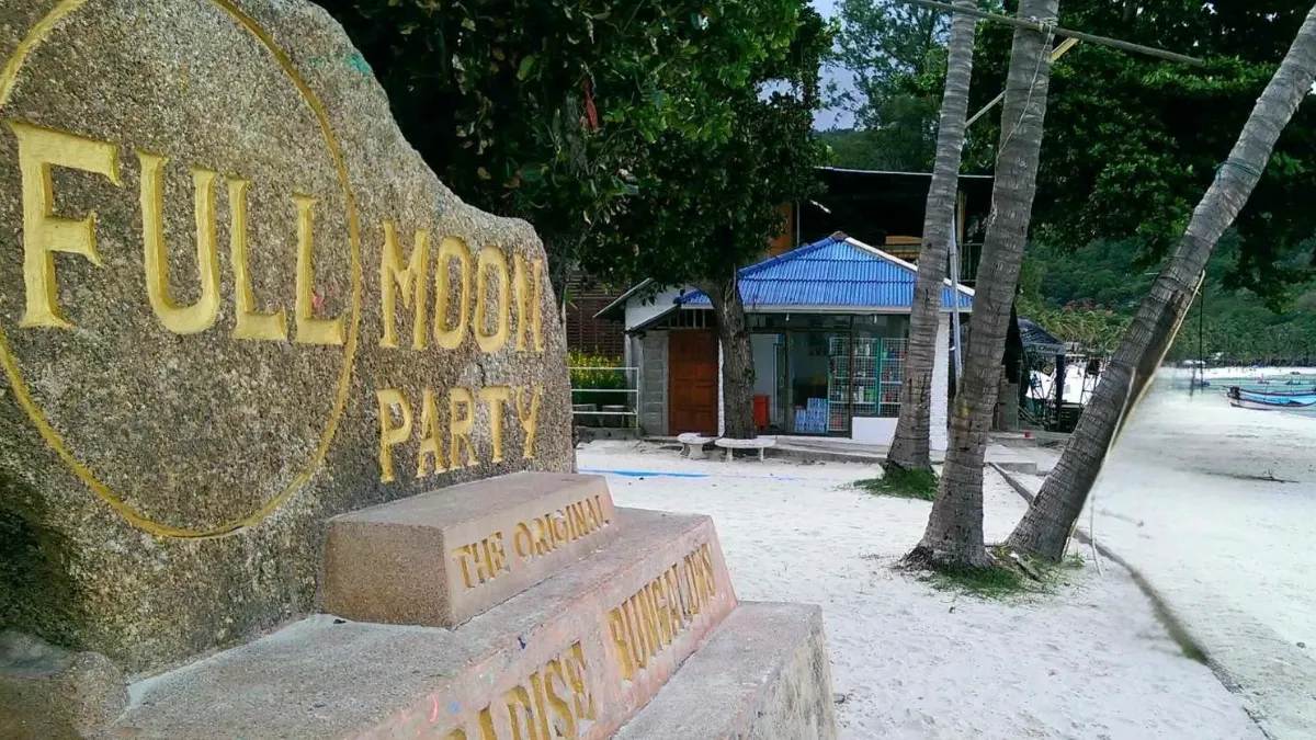 Stone sign for Full Moon Party on a beach with palm trees and a small building nearby