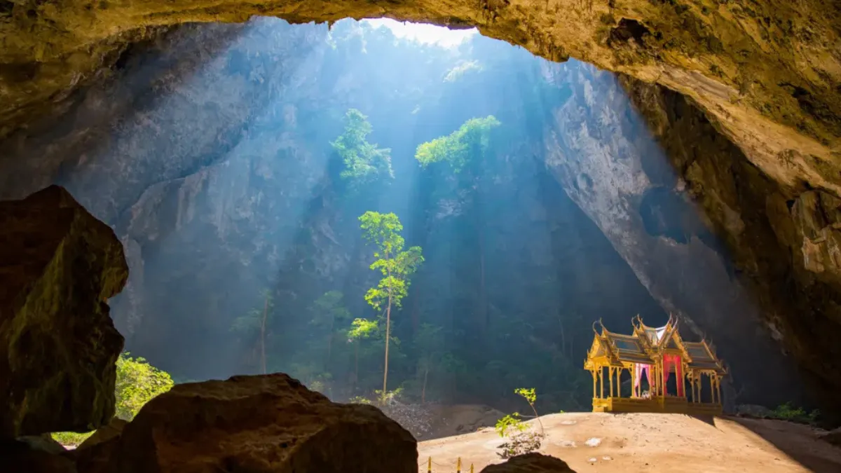 Sunlight streams into a large cave, illuminating a small ornate shrine on the cave floor
