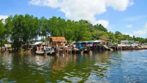 A riverside village with stilt houses, boats, and docks along a calm waterway under a blue sky