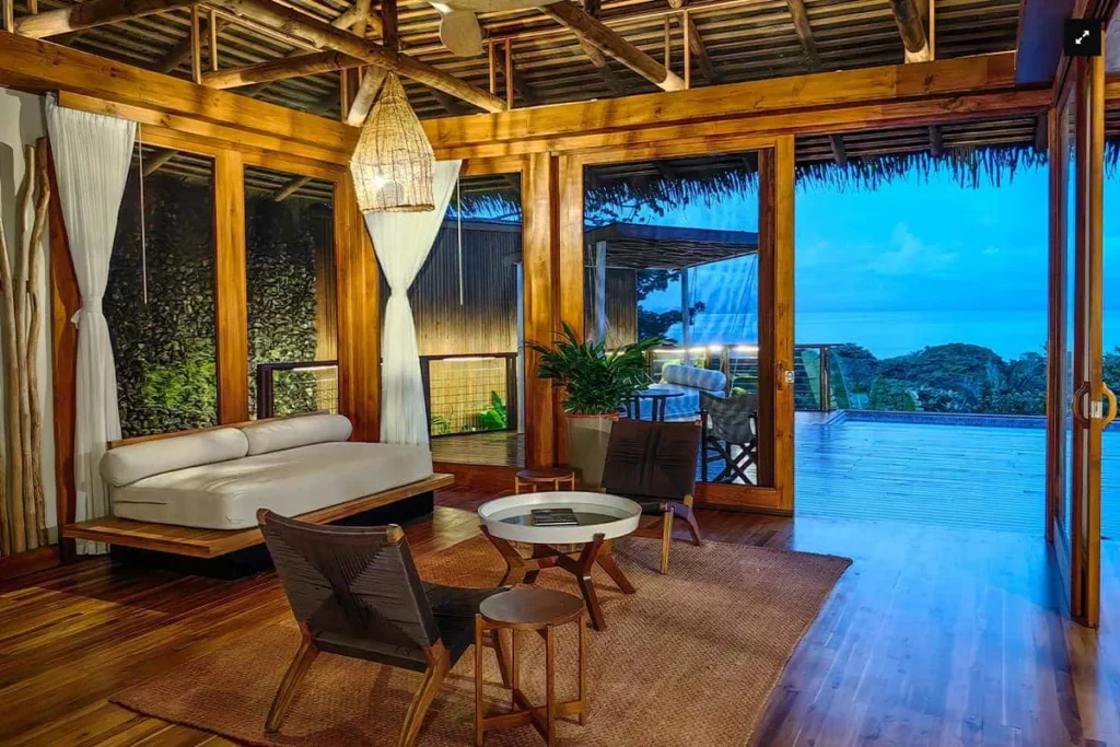 Open-air wooden lounge with a sofa, chairs, and a view of the ocean and deck