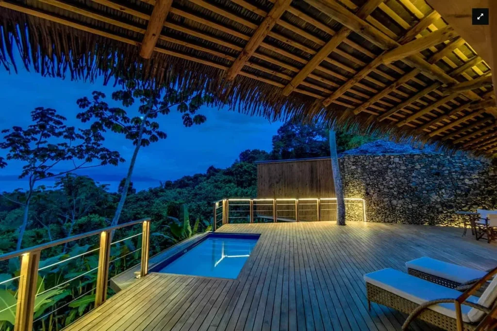 Covered wooden deck with a small lit pool overlooking a tropical forest at dusk