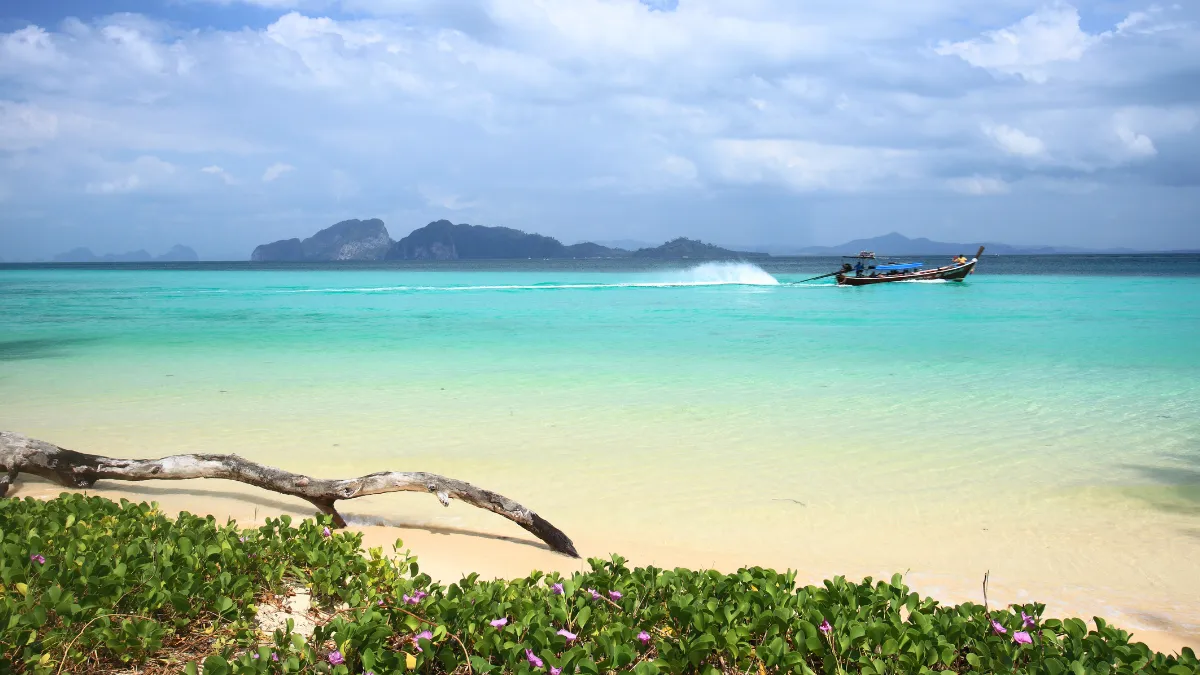 Tropical beach with turquoise water, a long-tail boat, and distant islands under a cloudy sky