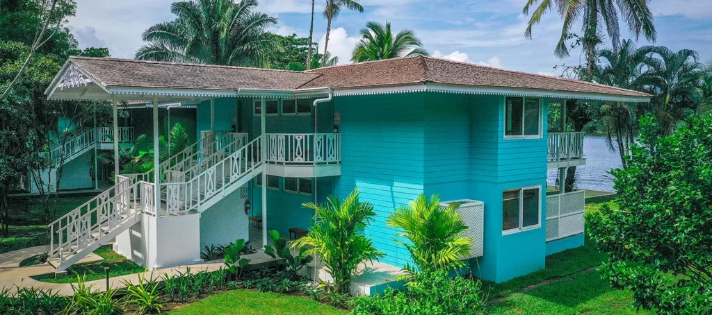 A turquoise tropical house with white stairs and balconies surrounded by palm trees near the water