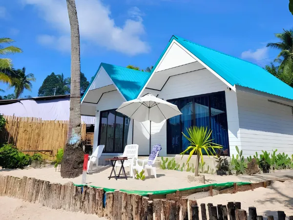 Beachfront cottages with turquoise roofs, patio chairs, and an umbrella under a blue sky