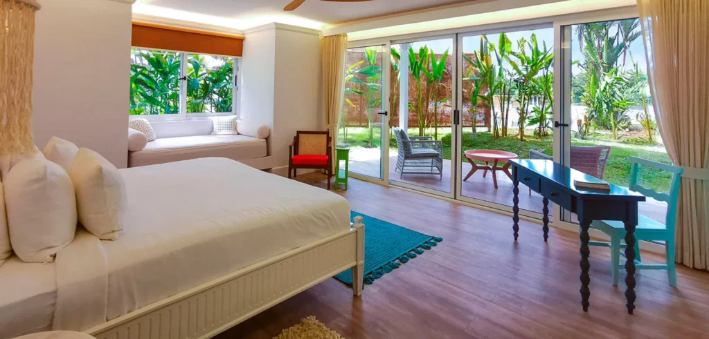 Bright bedroom with a bed, desk, and seating area opening onto a tropical patio through large glass doors