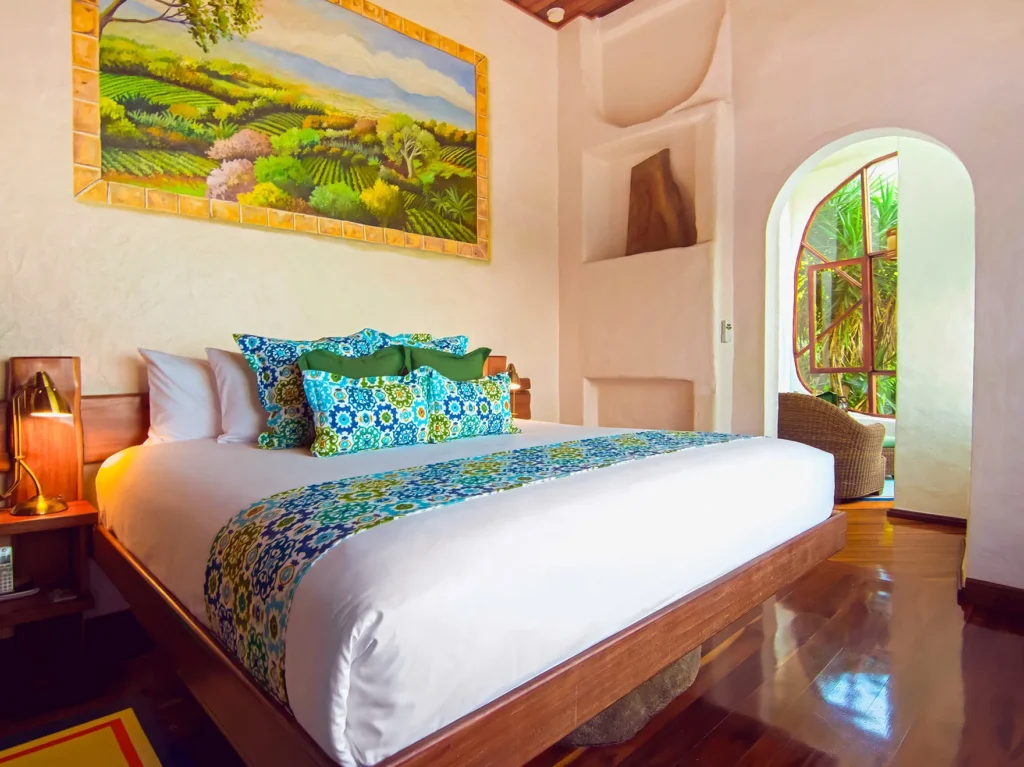 A bright bedroom with a large bed, colorful patterned pillows, and a landscape painting above the headboard