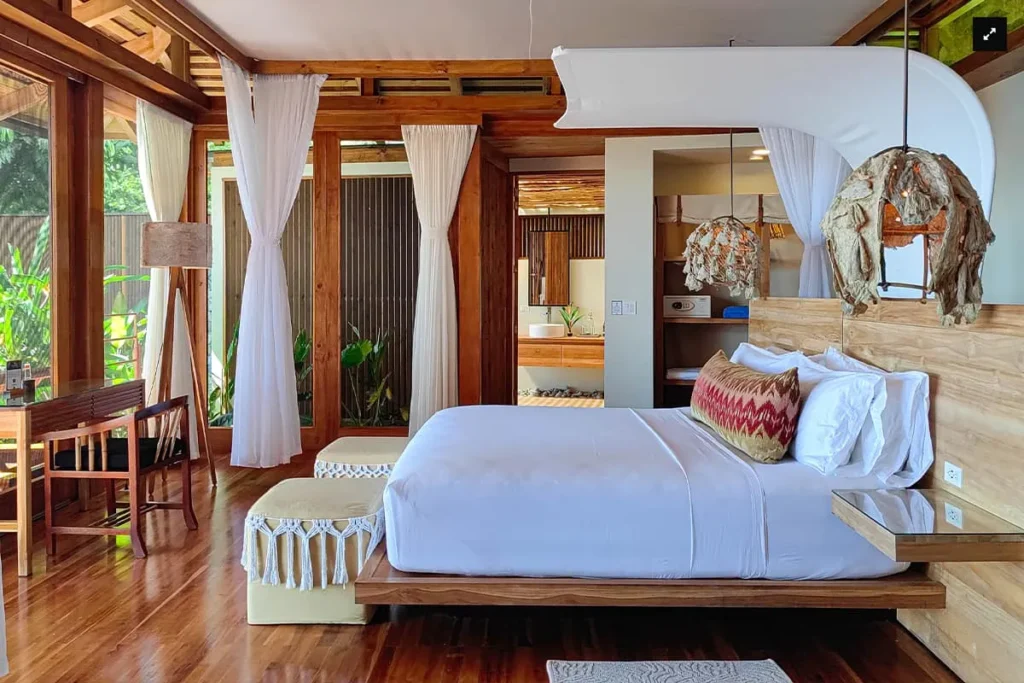 Modern bedroom with a wooden bed, white curtains, and large windows overlooking greenery