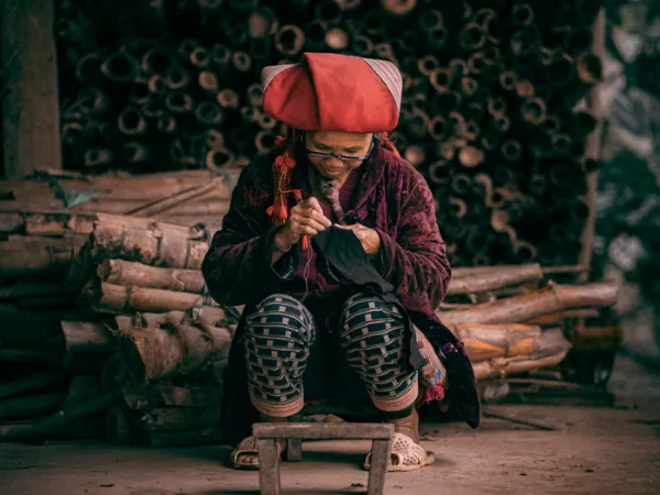 An elderly woman in traditional clothing sits and sews by hand indoors