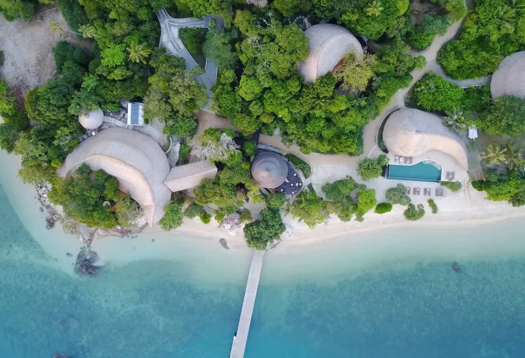 Aerial view of a tropical beachfront resort with thatched-roof villas, a pool, and a long pier over turquoise water