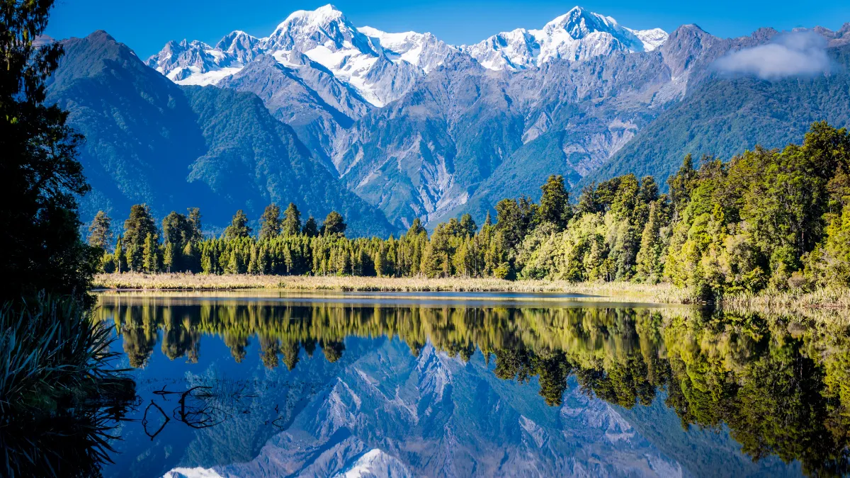 Snowy mountains reflected in a calm lake, with forested shores under a clear blue sky
