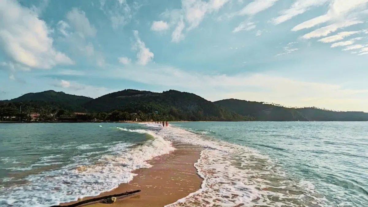 A sandbar extending into the sea with waves on both sides and mountains in the distance