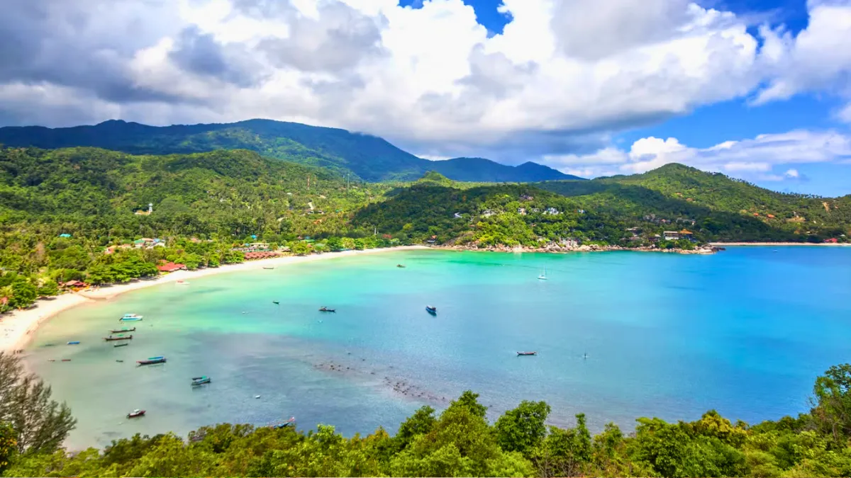 Tropical bay with turquoise water, sandy beach, boats, and green mountains under cloudy sky