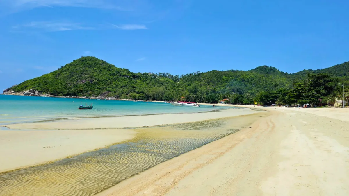 Tropical beach with turquoise water, a small boat, and green hills under a blue sky