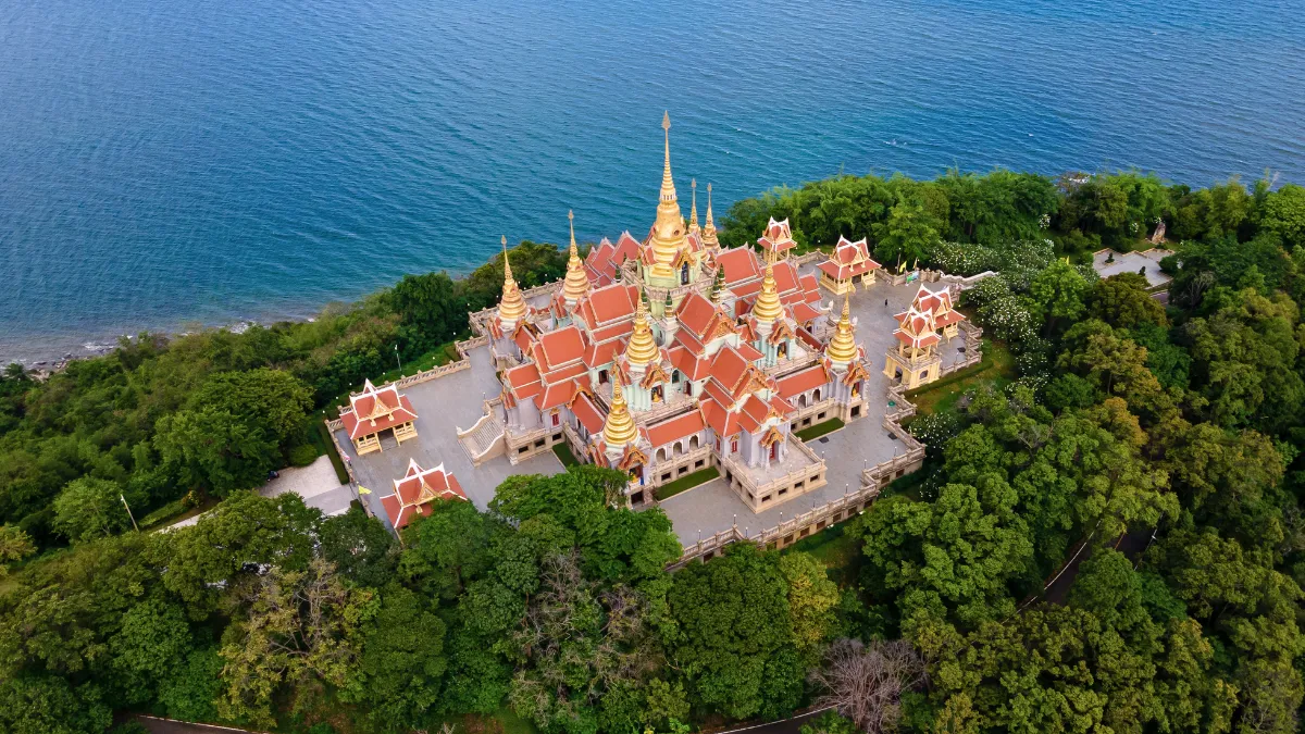 Aerial view of a Thai temple complex with golden spires beside the sea, surrounded by trees