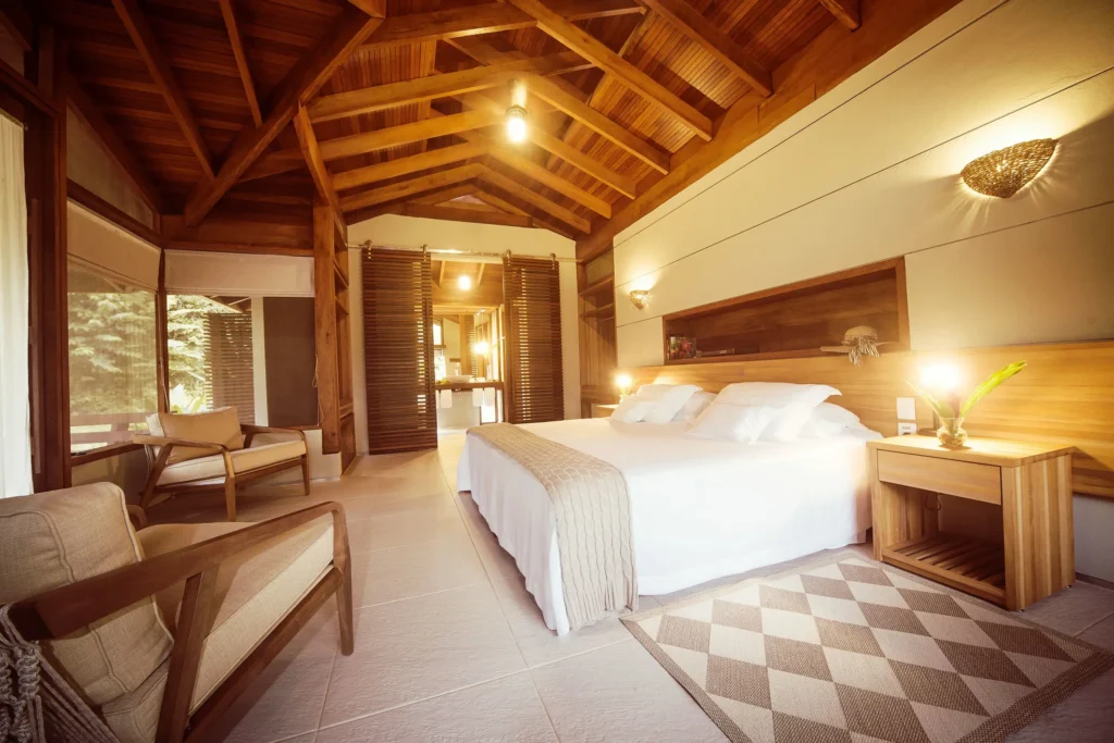 A warm wood-paneled bedroom with a king bed, lounge chairs, and large windows