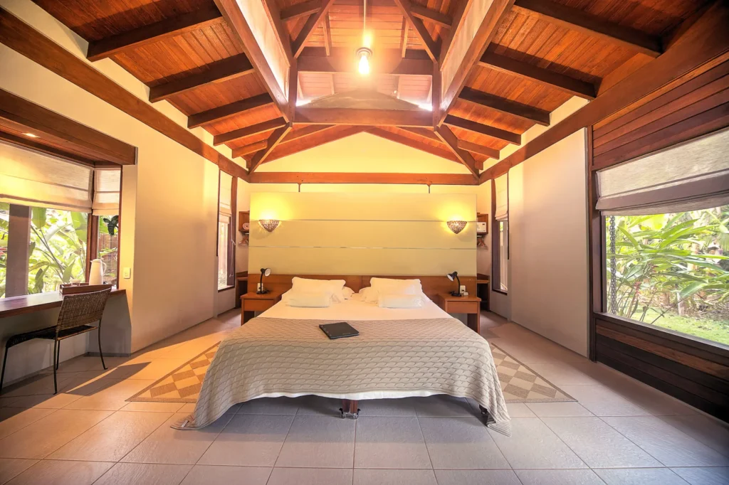A bright bedroom with a wooden vaulted ceiling, king bed, and large windows overlooking tropical greenery
