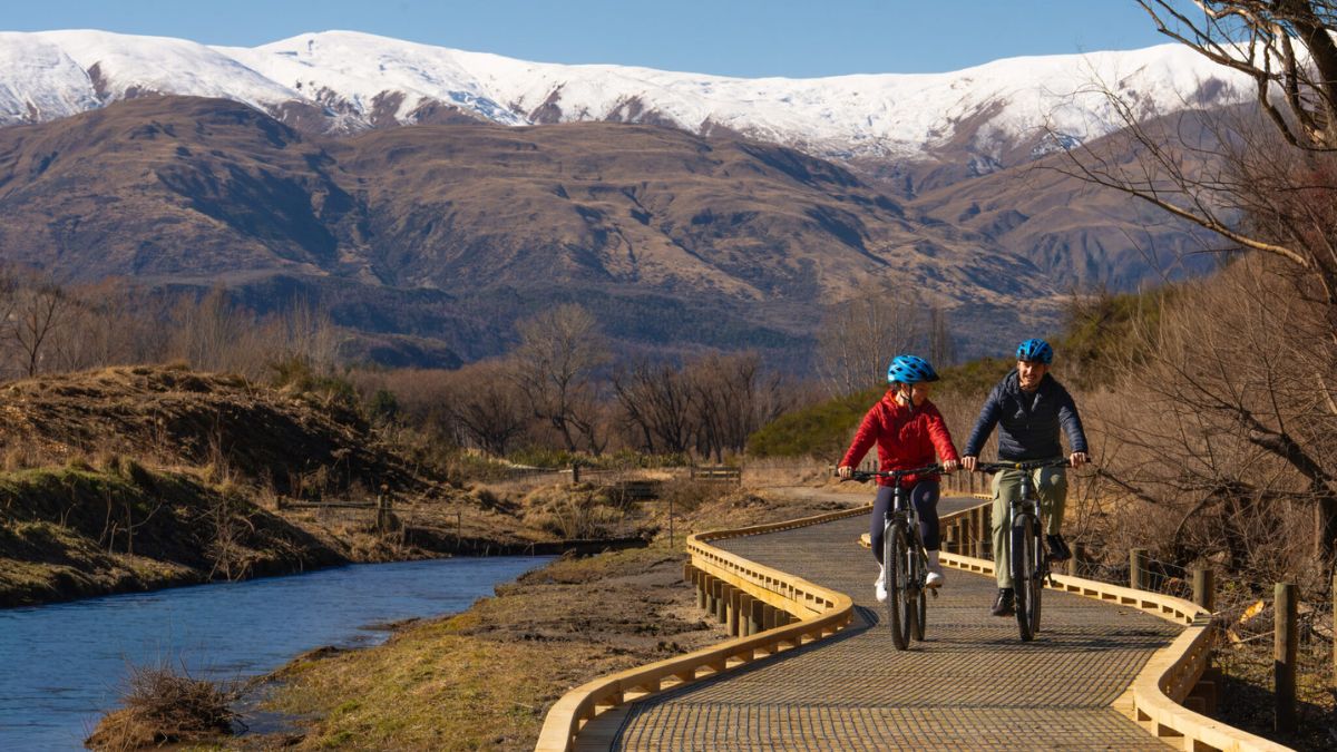 Two cyclists ride along a riverside boardwalk with snow-covered mountains in the background