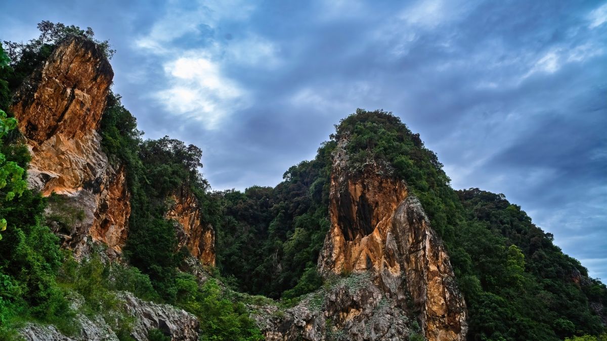Jagged rock cliffs covered in dense green vegetation beneath a cloudy blue sky