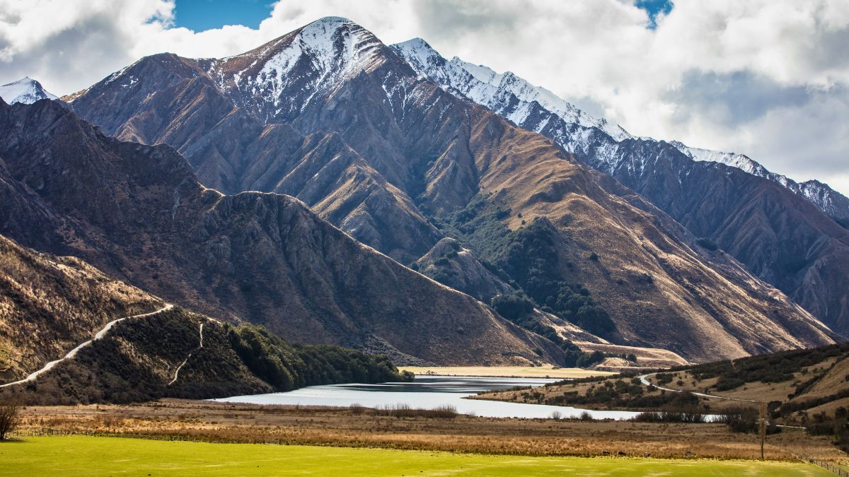 Snow-capped mountains rise above a lake and winding road, with golden hills and green fields in the foreground