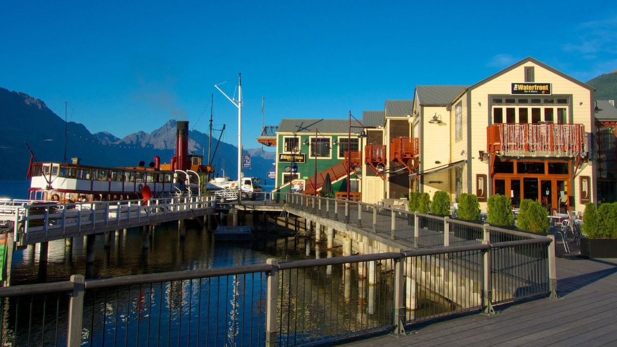 Waterfront marina with boats, boardwalk restaurants, and snow-capped mountains in the background
