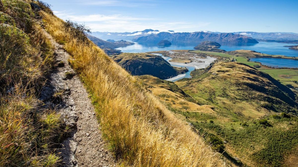A narrow trail winds along a grassy mountainside above a blue lake, rolling hills, and distant snow-capped mountains