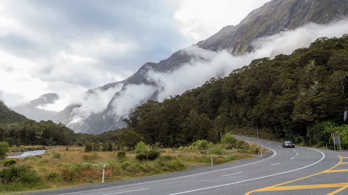 A winding road through forested mountains, with low clouds around the peaks and a stream beside the road