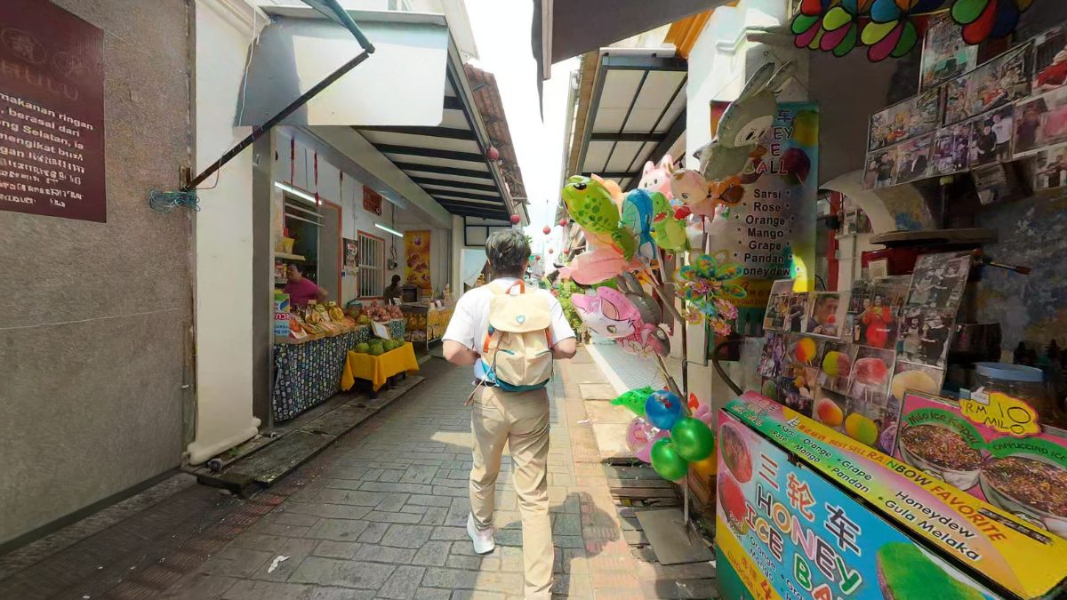 A person walks through a narrow market alley lined with food stalls, colorful balloons, souvenirs, and posters