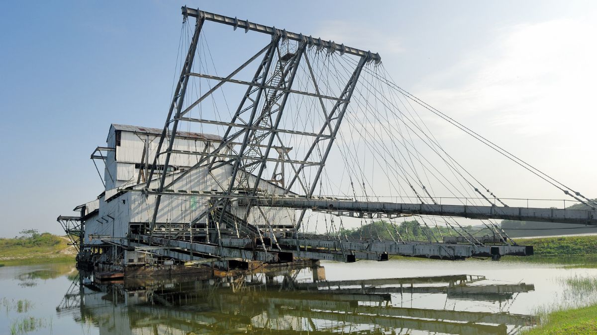 An abandoned industrial dredge with a long conveyor and suspended cables sits in a calm river