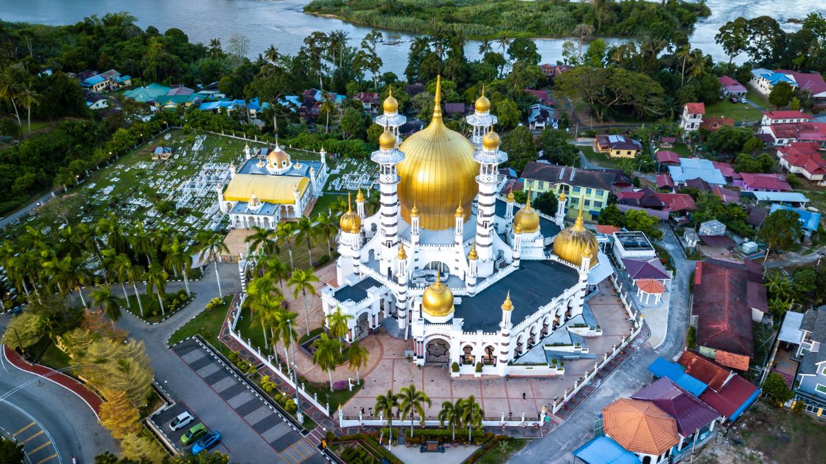 Aerial view of a grand mosque with golden domes, beside a cemetery and village near a river