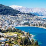 Panoramic view of a lakeside town surrounded by forested hills and snow-capped mountains - Queenstown travel guide