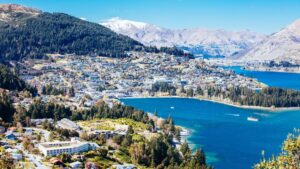 Panoramic view of a lakeside town surrounded by forested hills and snow-capped mountains - Queenstown travel guide