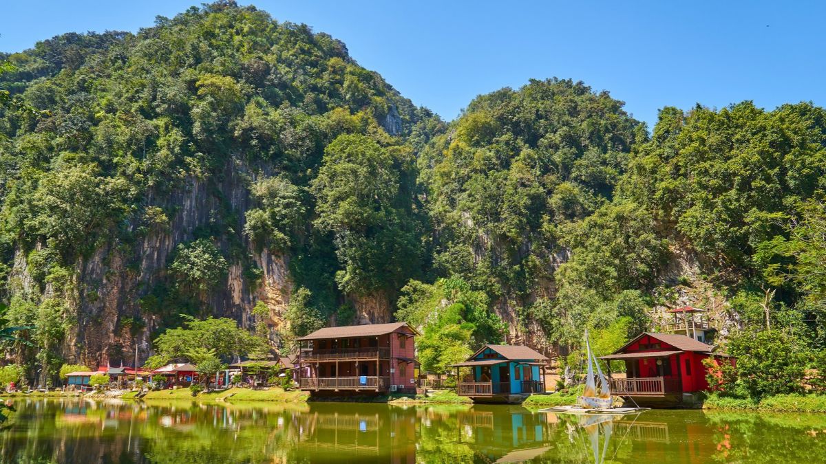 Lakeside village beneath lush, forested limestone cliffs, with colorful buildings and a small sailboat on the water - Ipoh Travel Guide